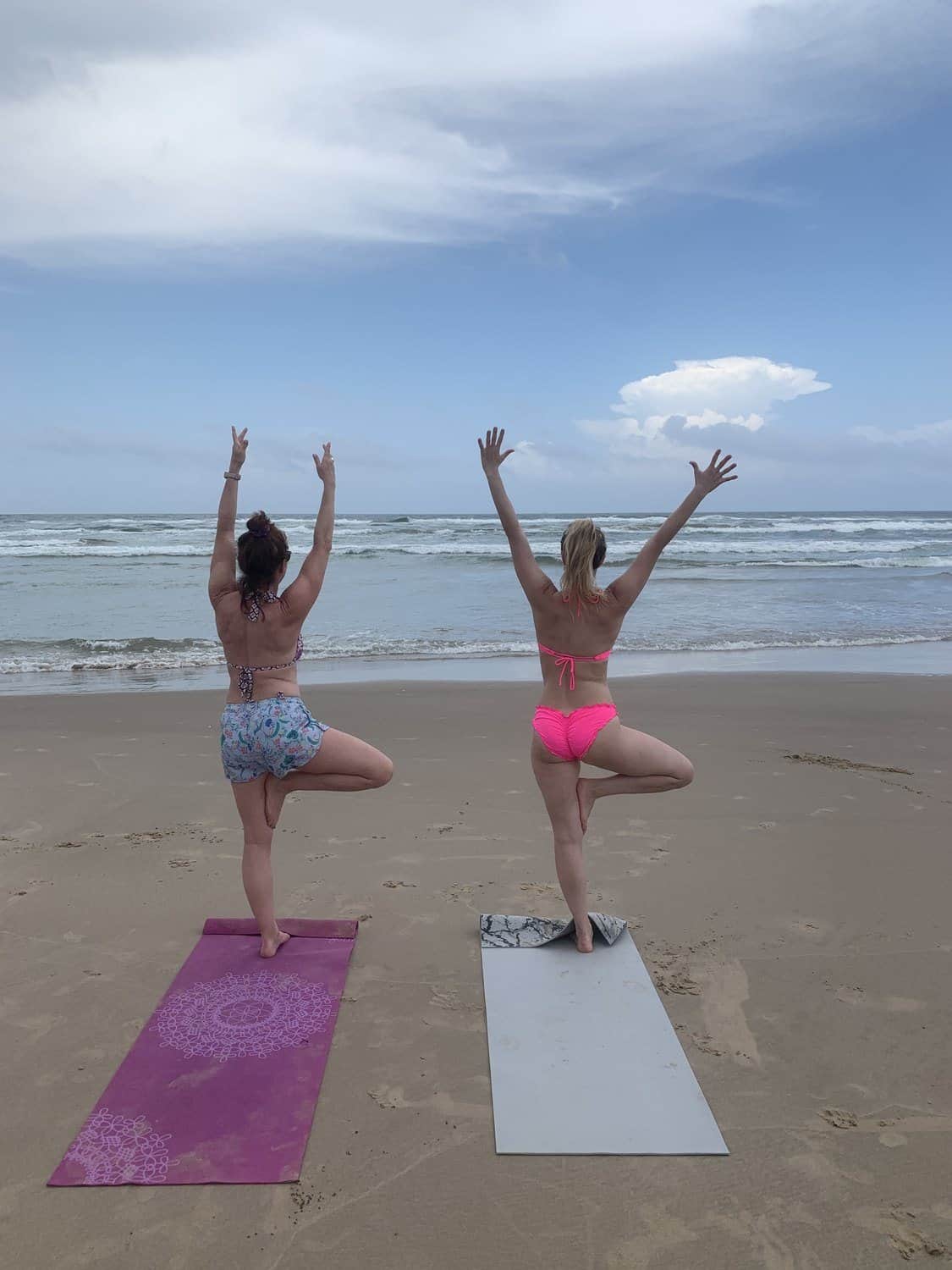 Two women balancing in tree pose on a beach at the water's edge